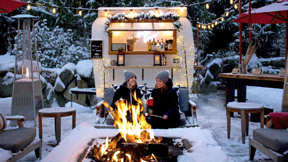 Winter firepit at Four Seasons Whistler with two women making s’mores beside a cozy outdoor drink trailer