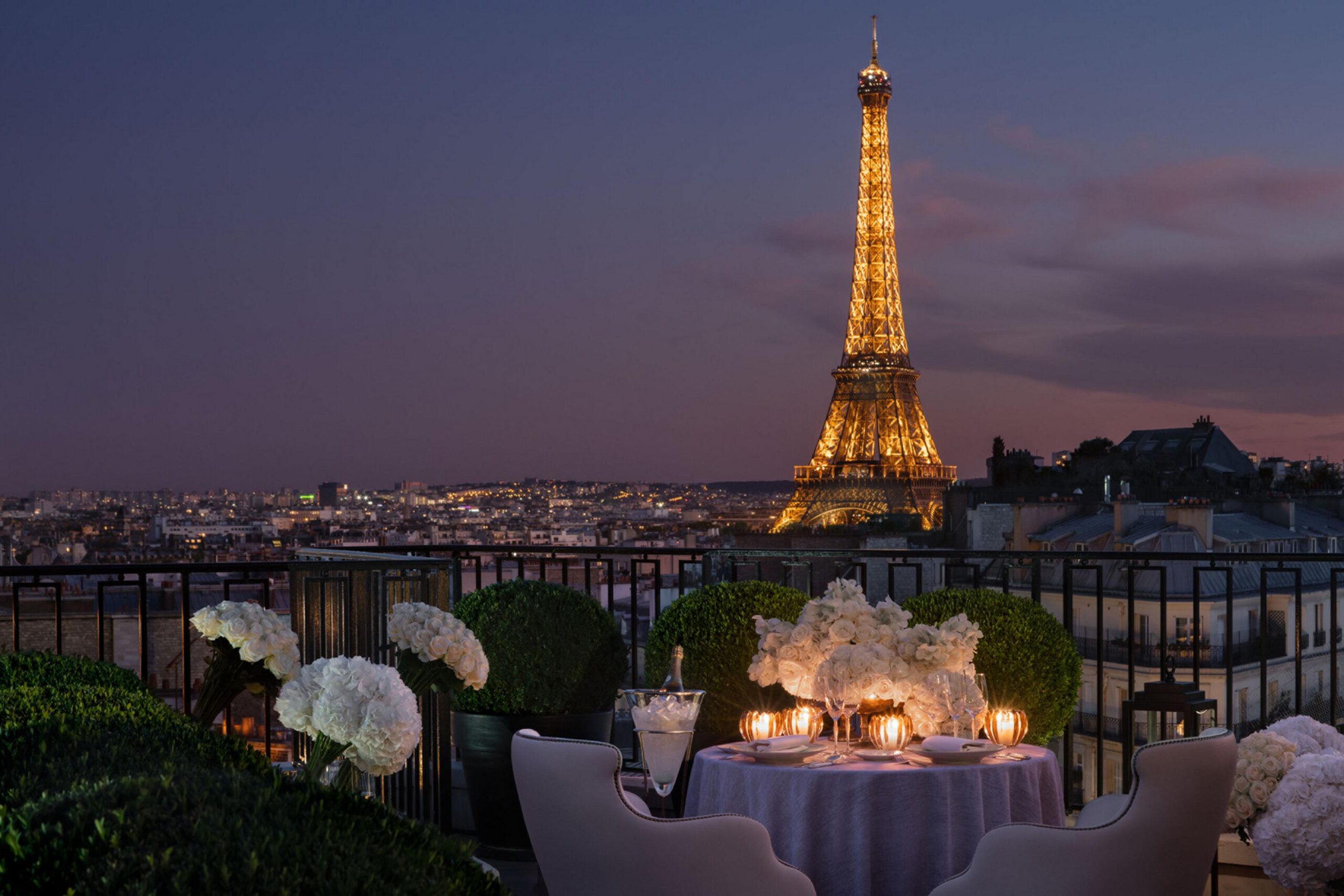 Night view from a Four Seasons George V Paris suite balcony with the Eiffel Tower illuminated in the background; a table set with white flowers, glowing candles, and a champagne bucket.