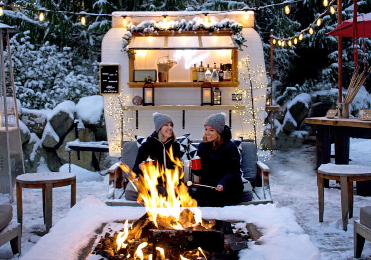 Two guests enjoying s'mores by outdoor fire pit with vintage camper serving hot chocolate at Four Seasons Whistler in winter snow